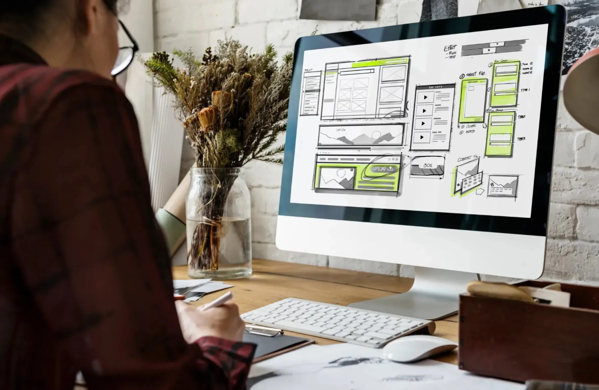 A web designer working on an e-commerce website layout on a desktop computer, sketching wireframes and interface designs at a creative workspace.