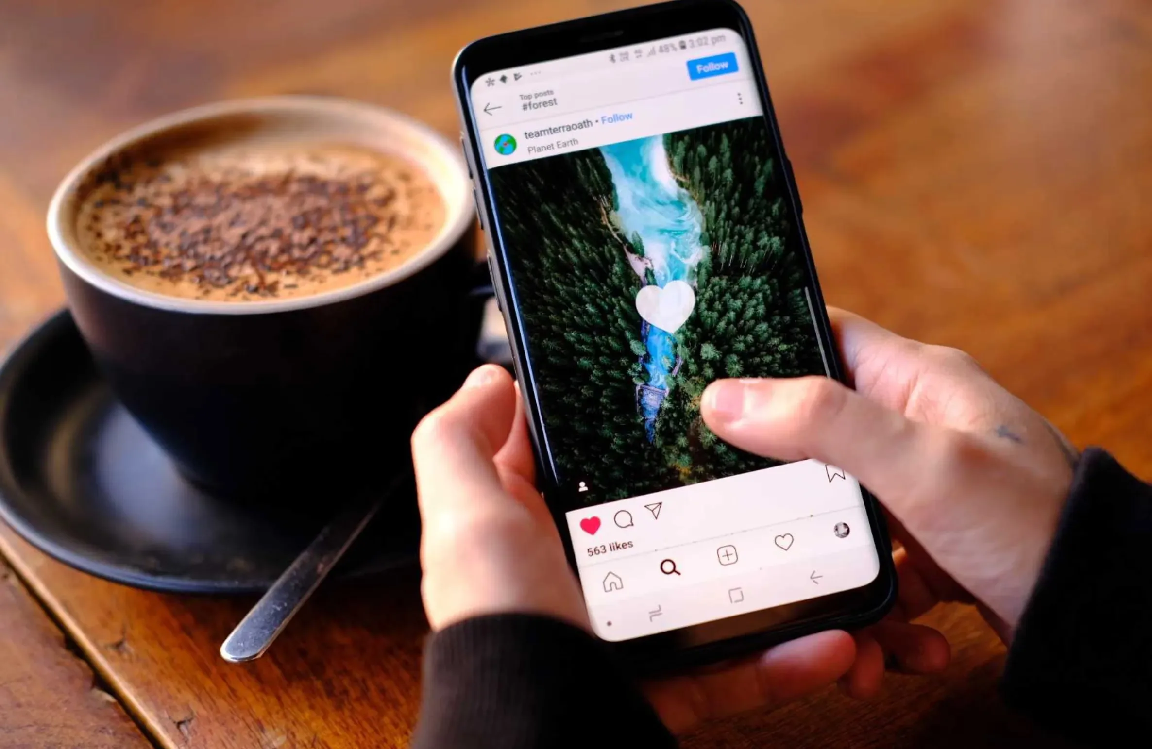A person holding a smartphone and scrolling through an Instagram post featuring a scenic aerial view, with a cup of coffee on the table beside them.